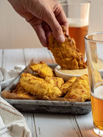 Final image of the gluten free chicken tenders. This image shows some beers that have been partially drunk, and a woman's hand dipping a chicken tender into some dipping sauce.