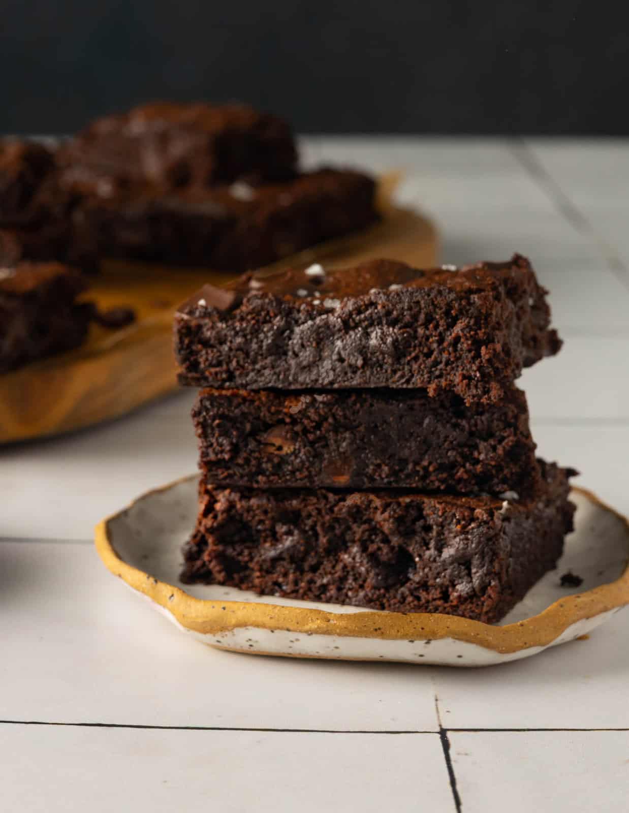 Final image of chocolate chunk brownies. There are three brownies stacked on a small dessert plate in front of a cutting board of brownies.