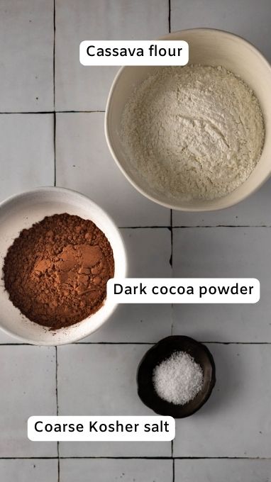 Ingredients for chocolate chunk brownies. There is a bowl with cassava flour, a bowl with dark chocolate cocoa powder, and a small bowl with kosher salt