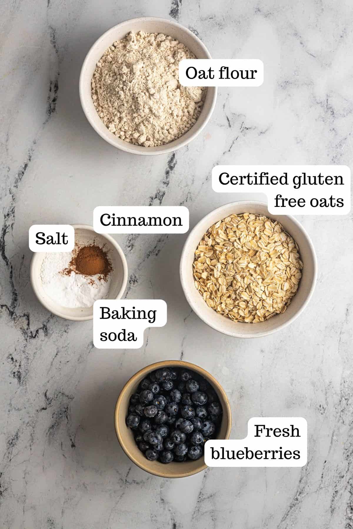 Image shows the dry ingredients for the Blueberry Banana Oat Muffins. In the bowls there is oat flour, certified gluten-free oats, cinnamon, salt, baking soda, and a bowl of fresh blueberries.