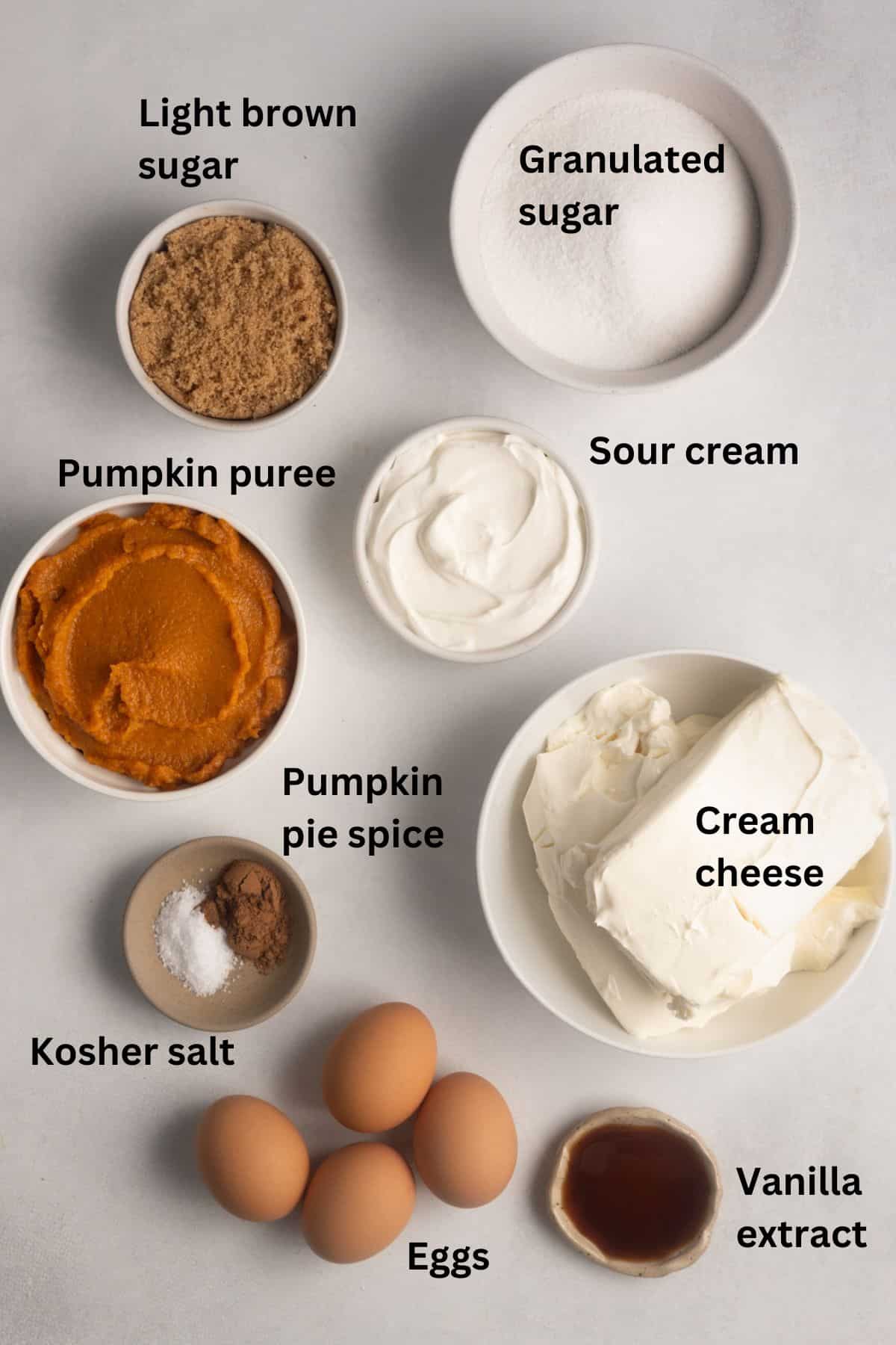 Image showing ingredients for gluten free pumpkin cheesecake. The ingredients shown are granulated sugar, light brown sugar, sour cream, pumpkin puree, cream cheese, pumpkin pie spice, Kosher salt, eggs, and vanilla extract.