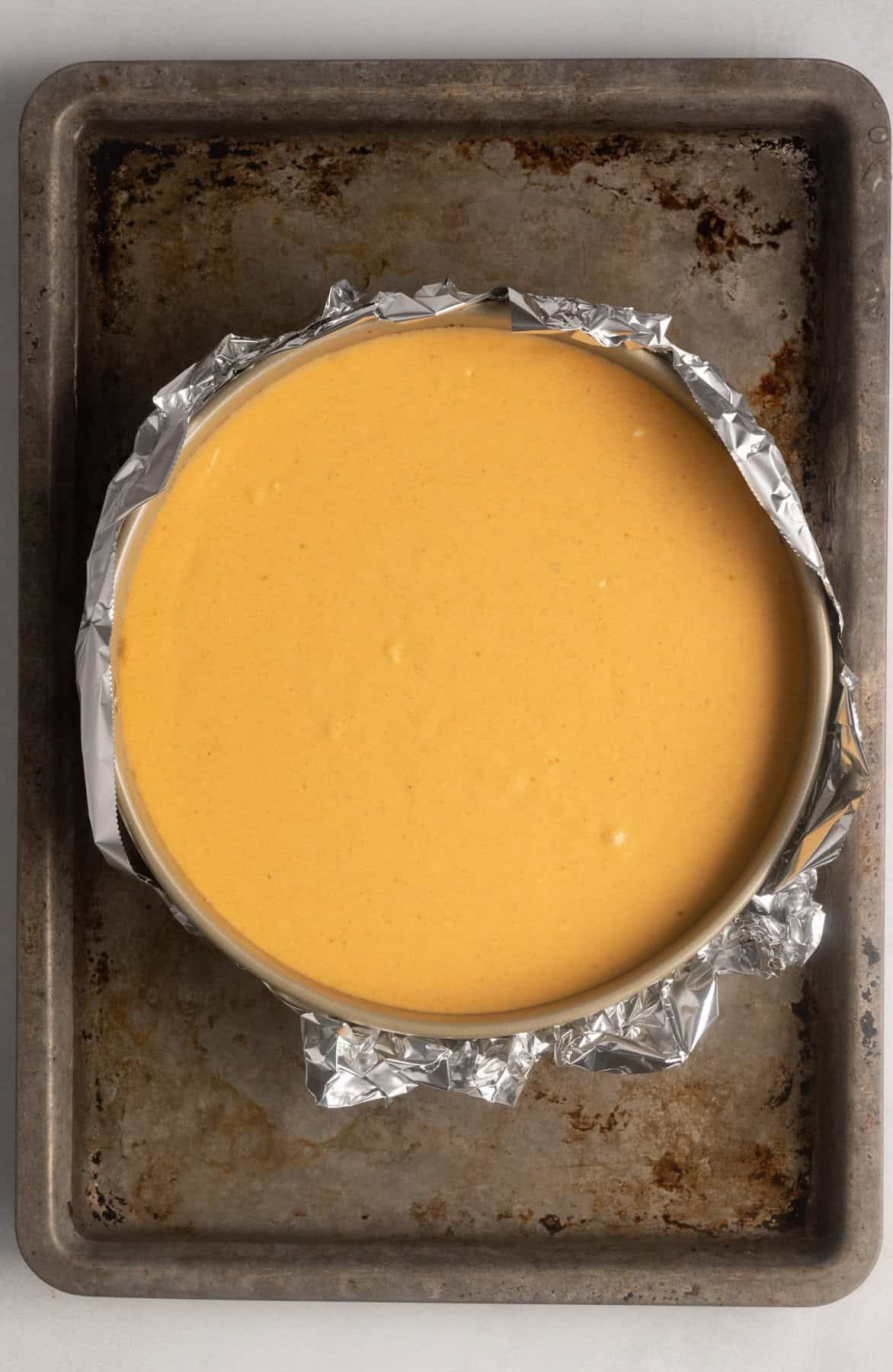 Gluten free pumpkin cheesecake has been added to the springform pan where the crust has been cooling. Water has been added to the sheet pan that the gluten free pumpkin cheesecake sits on. This forms a water bath that allows for more even baking.