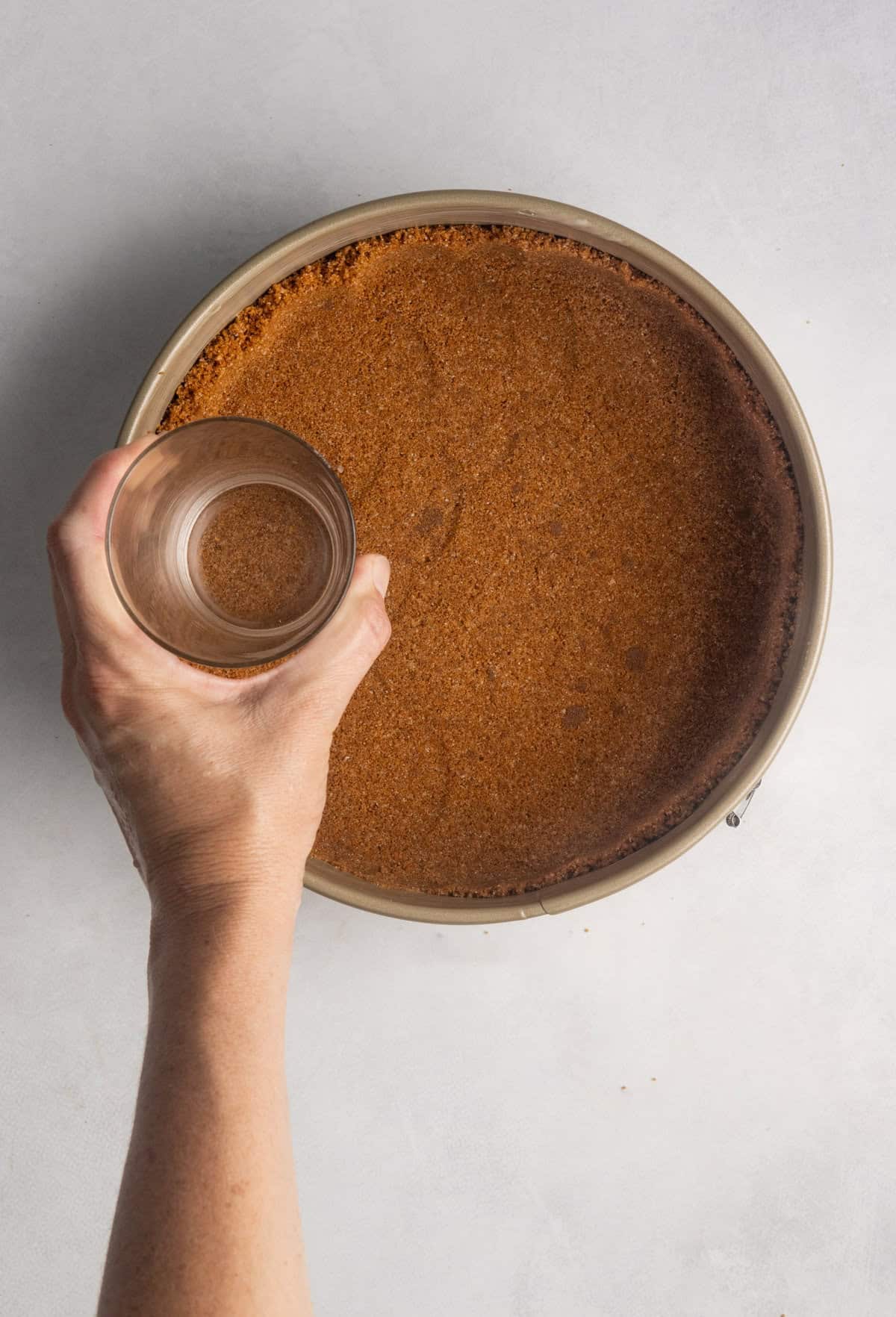 The crust for gluten free pumpkin cheesecake is being pressed into a lightly greased 8-inch springform pan. A glass is used to evenly compress the mixture.