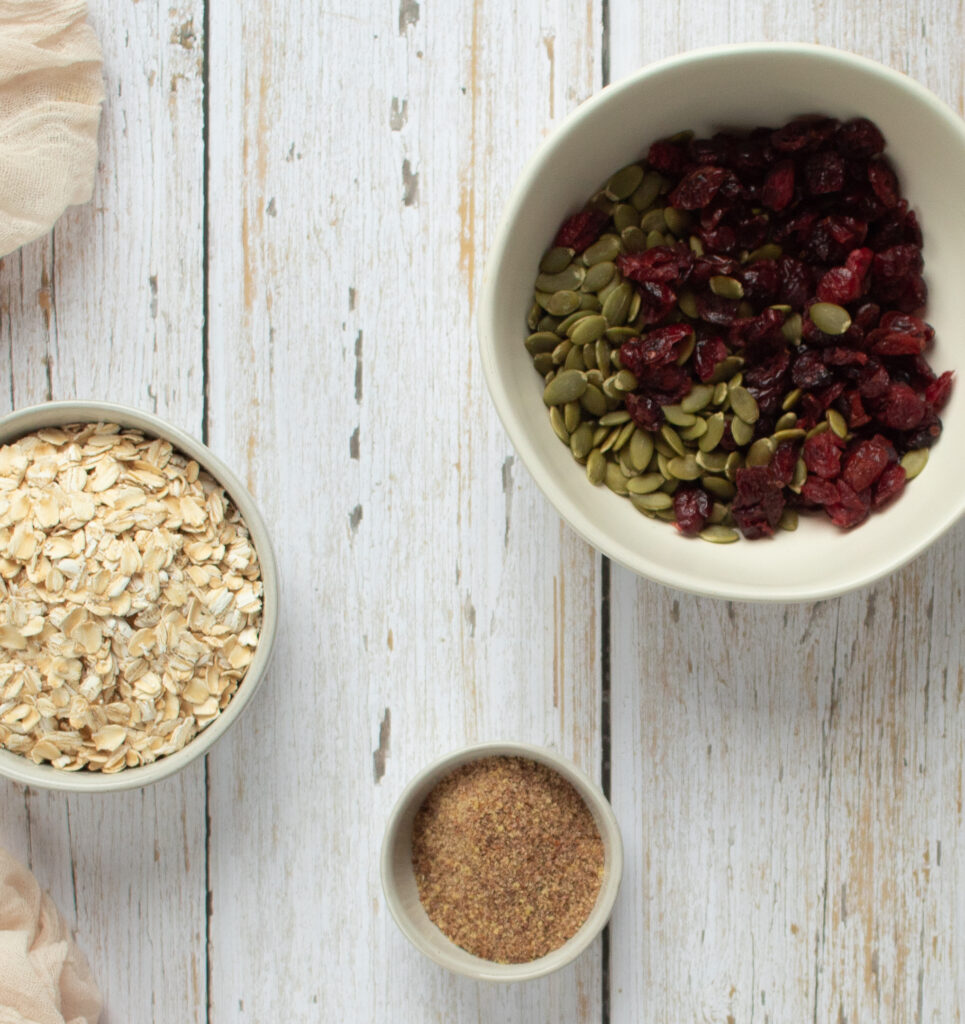 bowls of ingredients with cranberries, pumpkin seeds, flax meal, and oats